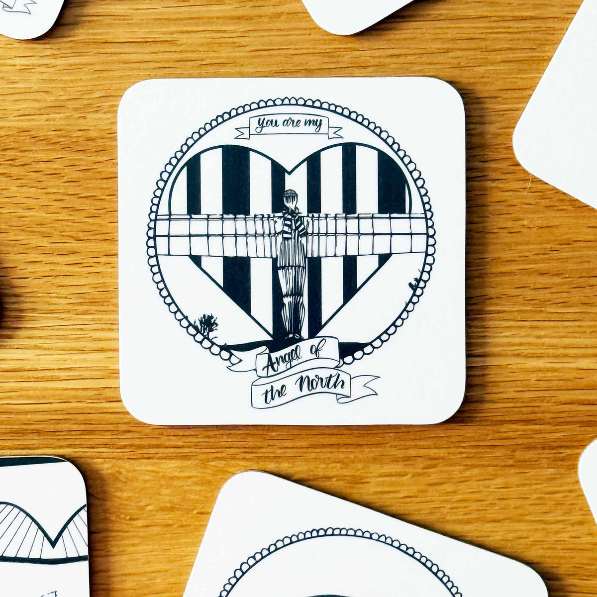 A square white coaster with rounded corners featuring a black and white graphic of the Angel of the North over a striped heart, with banners reading 'You are my Angel of the North', placed on a wooden tabletop with other coasters partially visible around it.