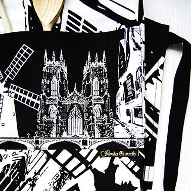 PowderButterfly York Apron with black and white illustration of York monuments and landmarks. Woman wears it while baking macaroons. 