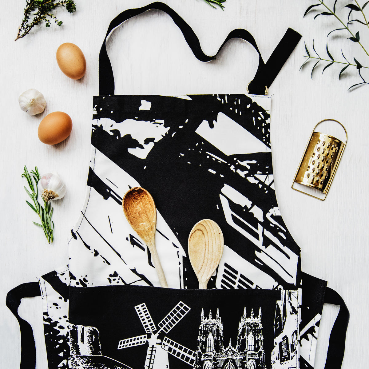 A black and white apron with a bold abstract York-themed pattern and a lower panel featuring white line drawings of Holgate windmills, York Minster and The Shambles, styled flat-lay on a white wooden surface with eggs, garlic, wooden spoons, a gold grater, and fresh herbs.