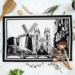 A rectangular tea towel or placemat with a black and white architectural illustration of landmarks in York, featuring  Holgate windmill. York Minster and the Shambles, styled on a white marble surface with a wooden spoon, copper utensils, and green herbs.