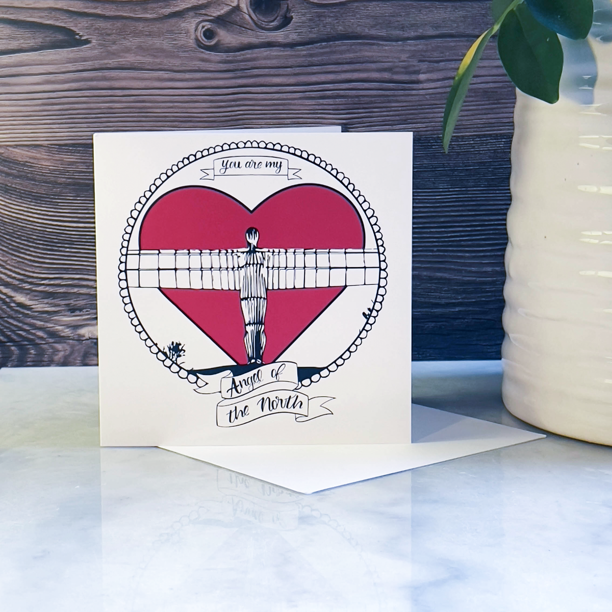 Greeting card with Angel of the North heart design and text on a marble surface with a wooden background next to a plant