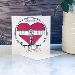 Greeting card with Angel of the North heart design and text on a marble surface with a wooden background next to a plant