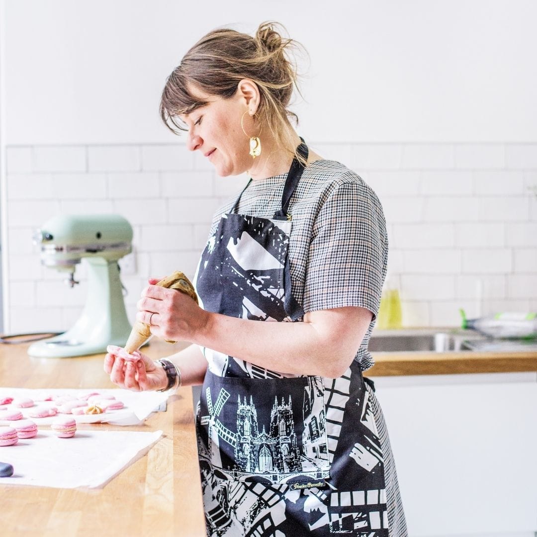 PowderButterfly York Apron with black and white illustration of York monuments and landmarks. Woman wears it while baking macaroons. 